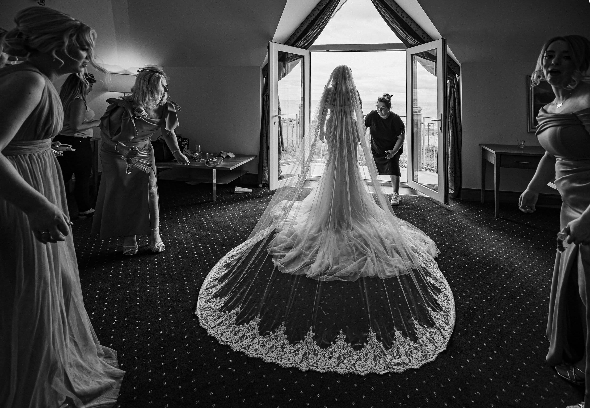 Bride standing in the window light at a Cork wedding venue as the bridesmaids run around fixing here