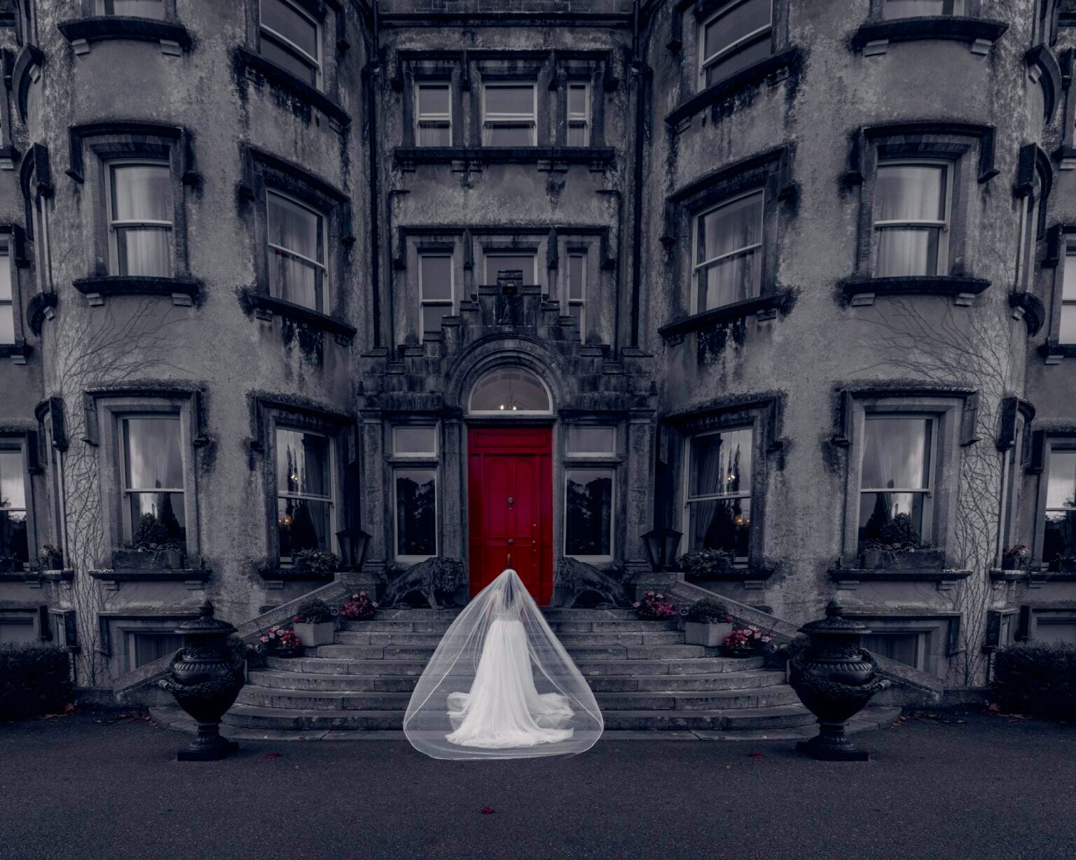 Black and white photograph of a bride in a long veil walking up the steps to the red door of a grand Irish castle