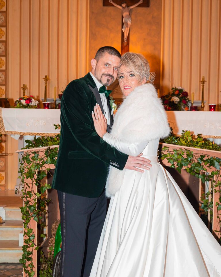 Bride in a white fur stole and groom in a green velvet jacket looking at the camera in front of a church altar decorated with ivy