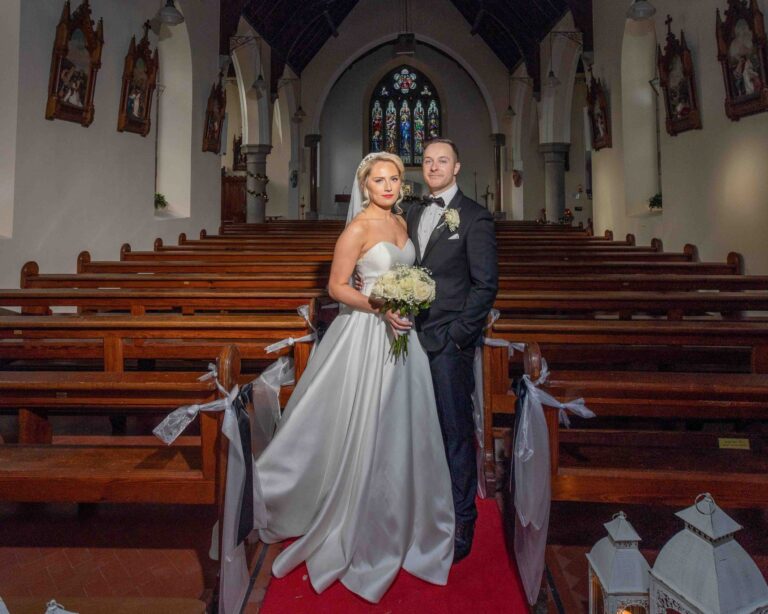 Bride and groom standing together and looking at the camera on the red carpet in the aisle of a church, white ribbons on the pews and stained glass behind them