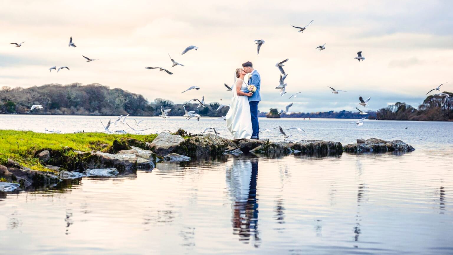 Bride and groom kissing at the edge of a lake in Killarney as seagulls take flight, wedding photography in Co. Kerry