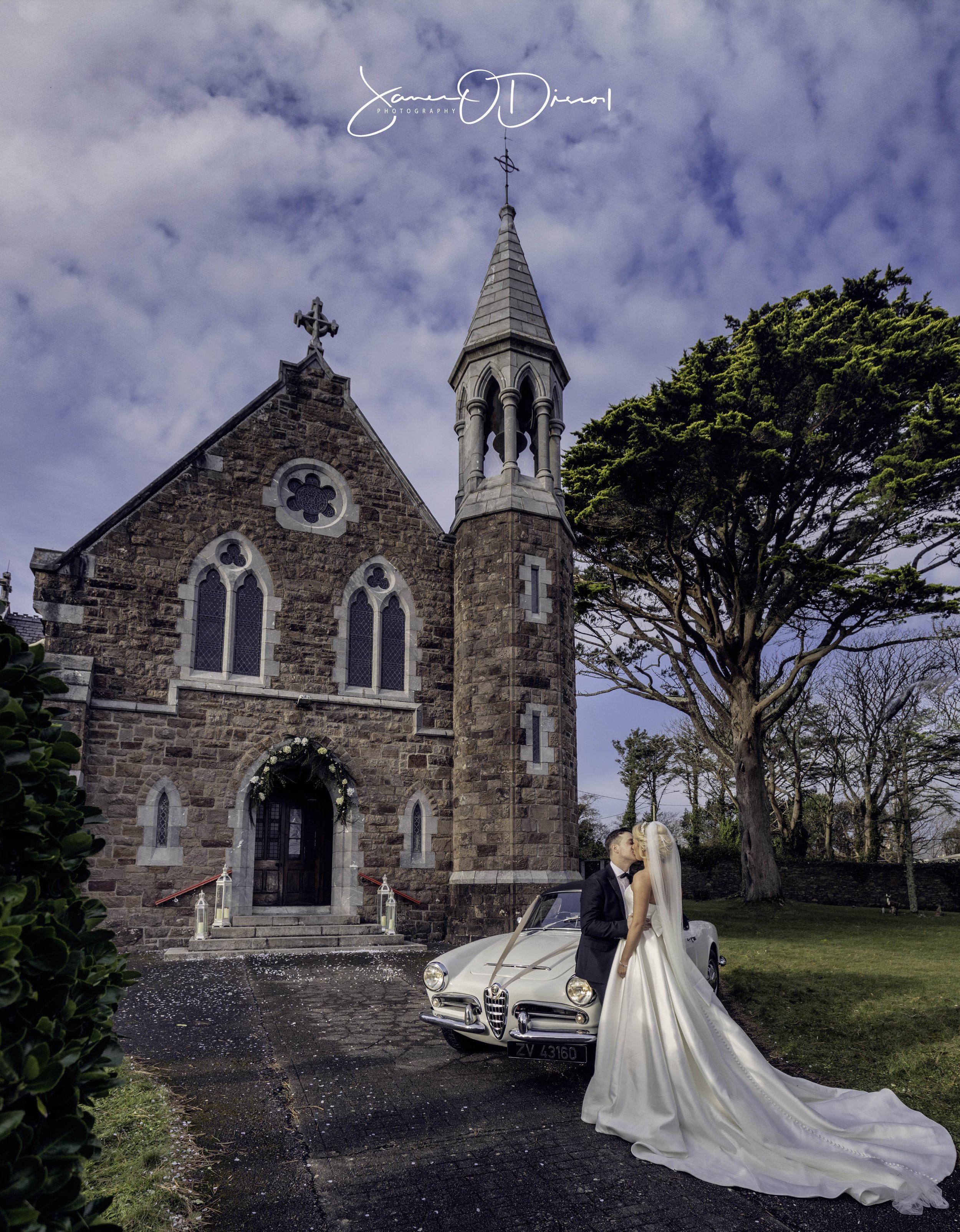 Posing next to a vintage car is a bride and groom outside a church on Valentia Island Co Kerry