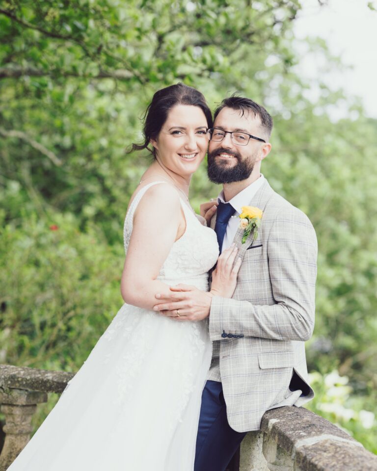Smiling bride and groom looking at the camera cheek to cheek beside a stone wall with green trees behind them