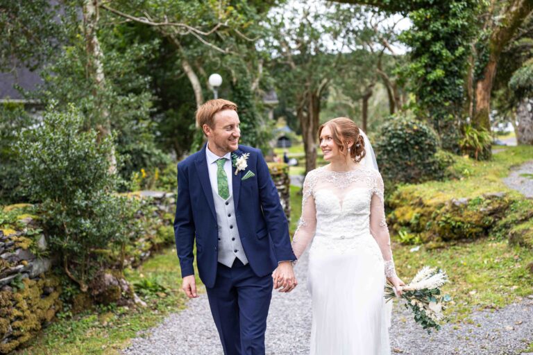 Groom and bride walking hand in hand along a gravel path between mossy stone walls, smiling at each other, Gougane Barra