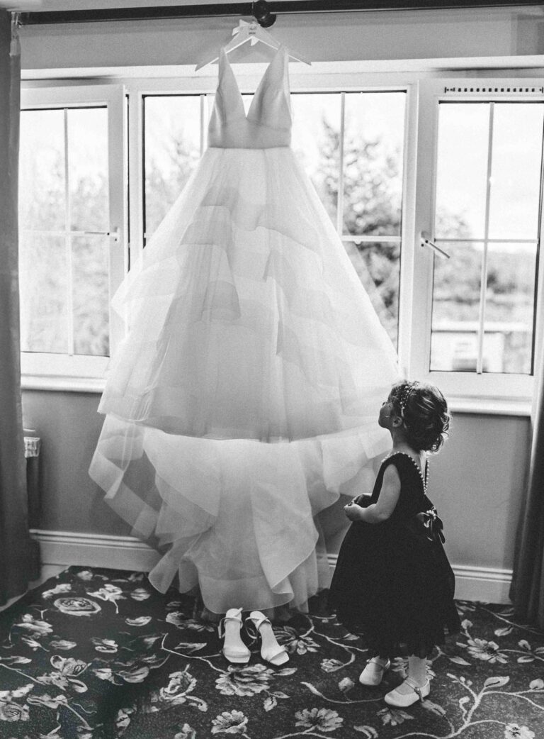 Black and white photo of a little girl looking up at a wedding dress hanging in the window on the wedding morning