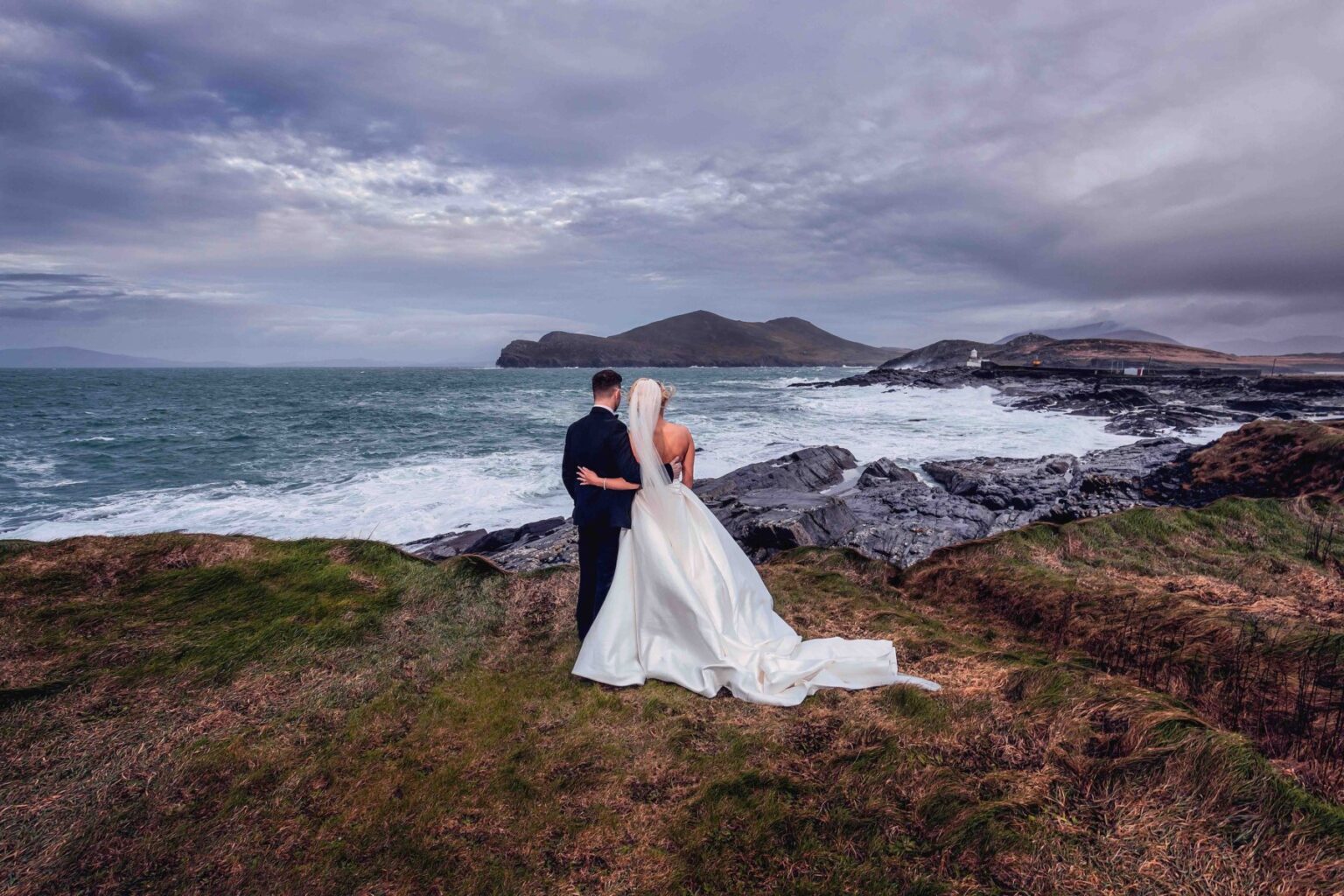 Bride and groom with their arms around each other looking out over stormy seas and rocky cliffs on the Wild Atlantic Way at Valentia Island, Co. Kerry