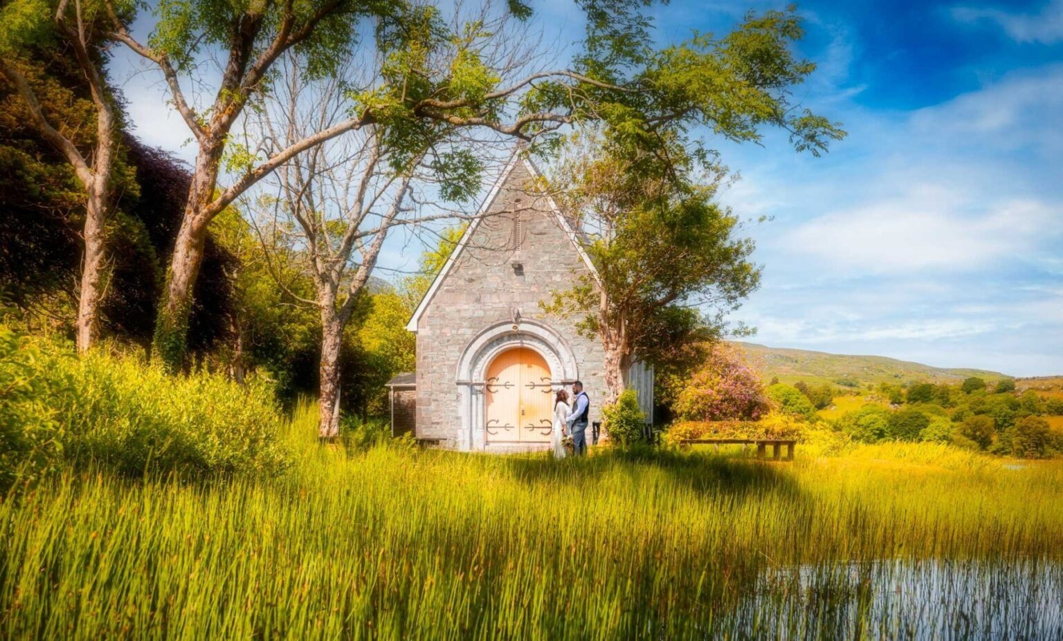 Bride and groom standing close in front of the little stone church at Gougane Barra, West Cork, surrounded by trees and long golden grass beside the lake