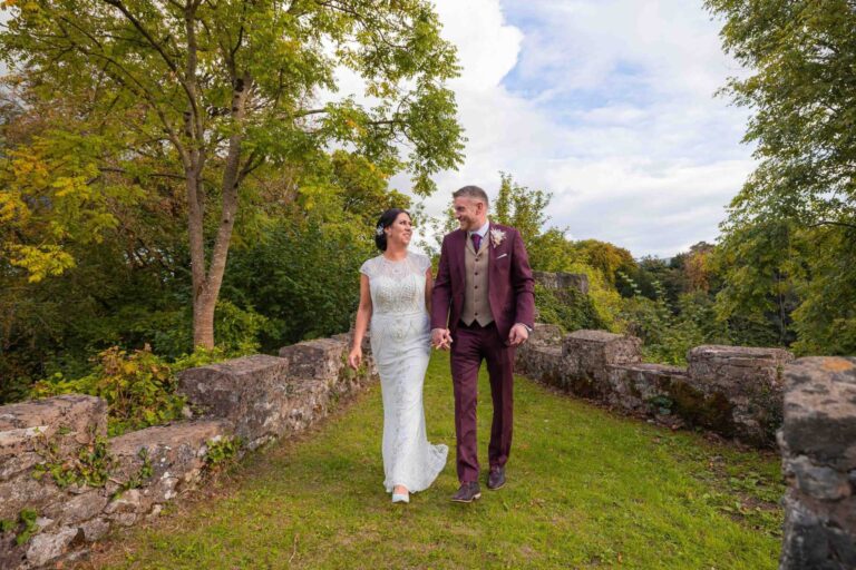 Bride and groom in a burgundy suit walking along a mossy stone wall in a garden
