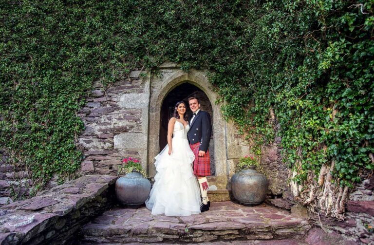 Bride and groom looking at the camera from inside a stone arched doorway covered in ivy, the groom in tartan