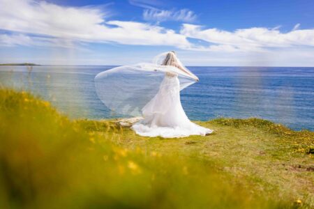 Wide framing of a bride with a long flowing veil standing on beach boulders with the groom at Long Strand in West Cork