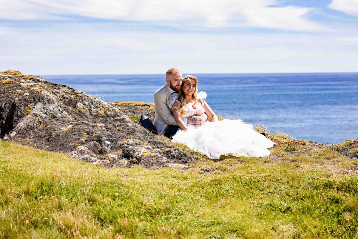 A bride and groom sitting together on a rugged, grassy cliffside overlooking the ocean during a wedding portrait session near Rosscarbery