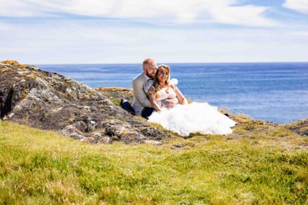 A bride and groom sitting together on a rugged, grassy cliffside overlooking the ocean during a wedding portrait session near Rosscarbery