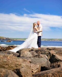 Full-length wedding photo of a bride and groom standing on dramatic beach rocks with a bright blue sky and coastline in West Cork