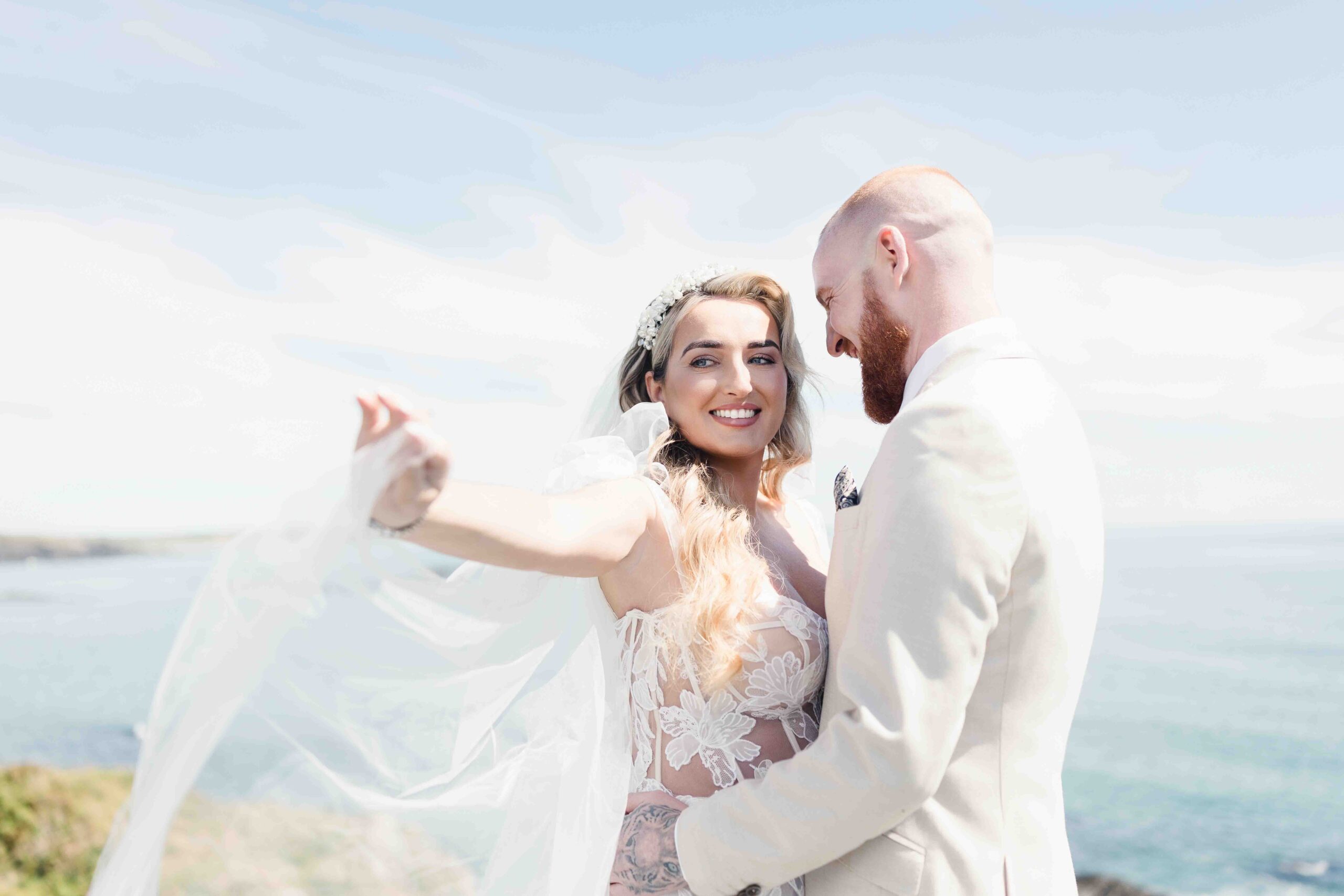 Bright and airy portrait of a bride looking over her shoulder with her veil blowing in the wind against a deep blue ocean background in West Cork