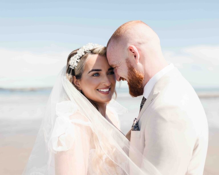 Bright, sun-drenched close-up portrait of a bride and groom standing together against a sunny beach background in Ireland