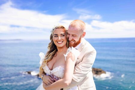 Full-length wedding photo of a bride and groom standing on dramatic beach rocks with a bright blue sky and coastline in West Cork