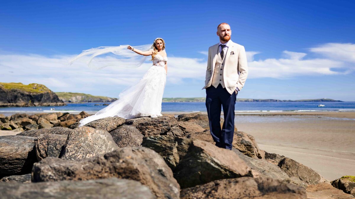 High-contrast, bright wedding portrait of a groom smiling at his bride while her veil catches the wind along the West Cork coast