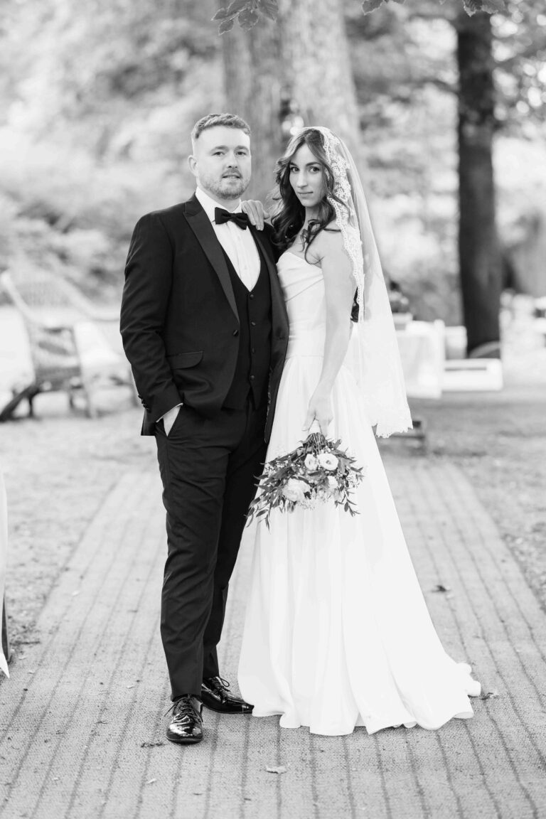 Black and white portrait of a groom in a tuxedo and a bride in a lace veil looking at the camera on a gravel path at Ballyvolane House, Cork