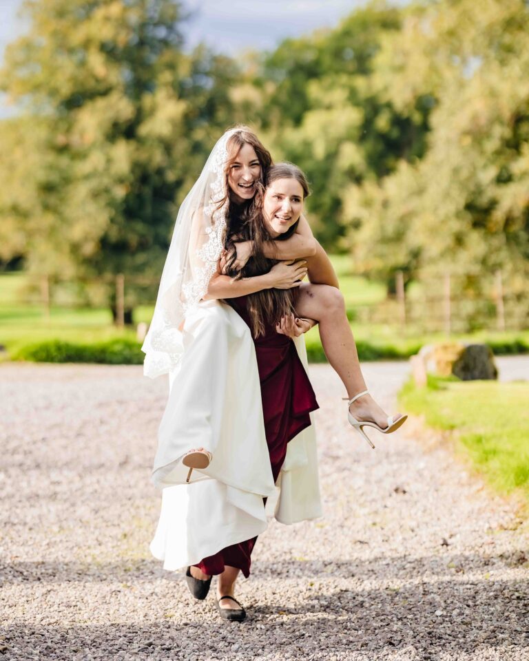 Bride laughing as she carries a bridesmaid on her back along the gravel at Ballyvolane House, Co. Cork