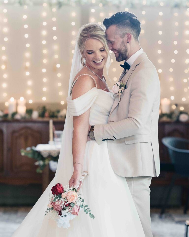 Bride and groom laughing and holding each other in front of fairy lights at The Village Barn, the bride holding her bouquet and veil
