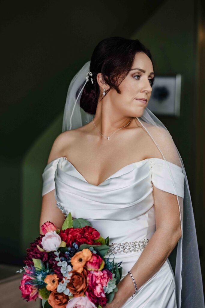 Bride glancing to the side in her veil and off-the-shoulder gown, holding a vibrant orange and pink bouquet