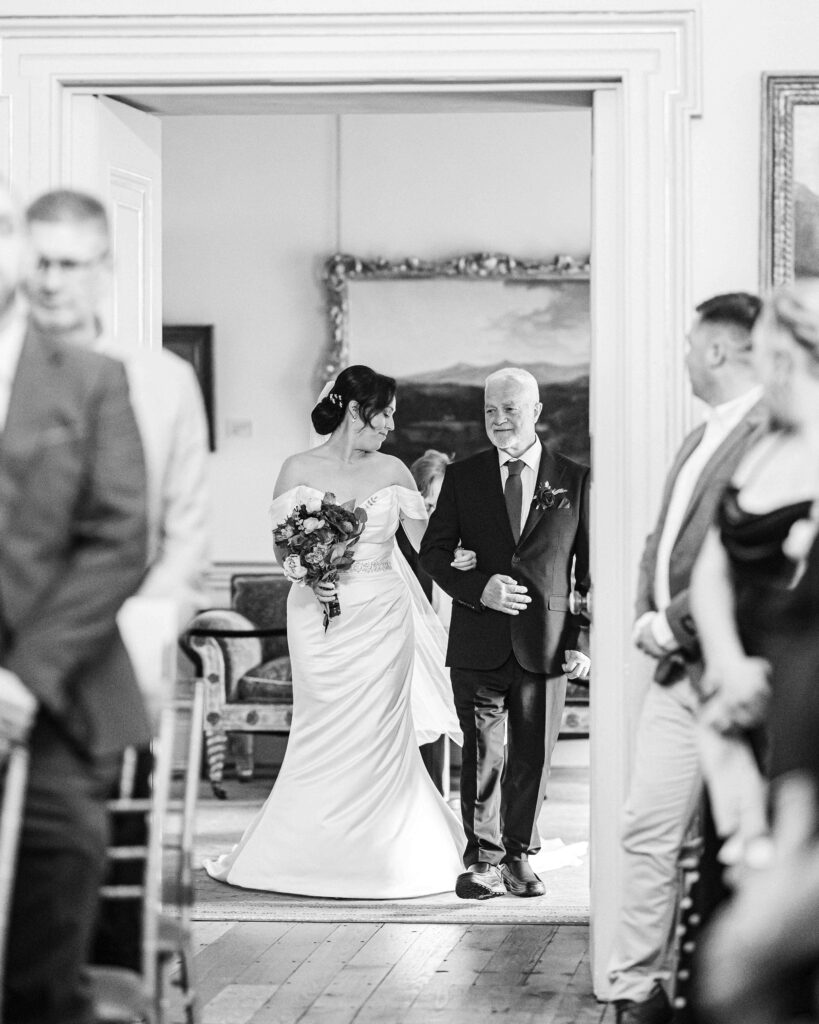 Black and white photo of the bride walking in on her father's arm through a doorway before the ceremony, Fota House, Cork