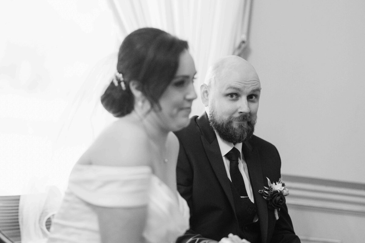 Black and white photo of the bride and groom sitting together during the ceremony as the groom glances at the camera