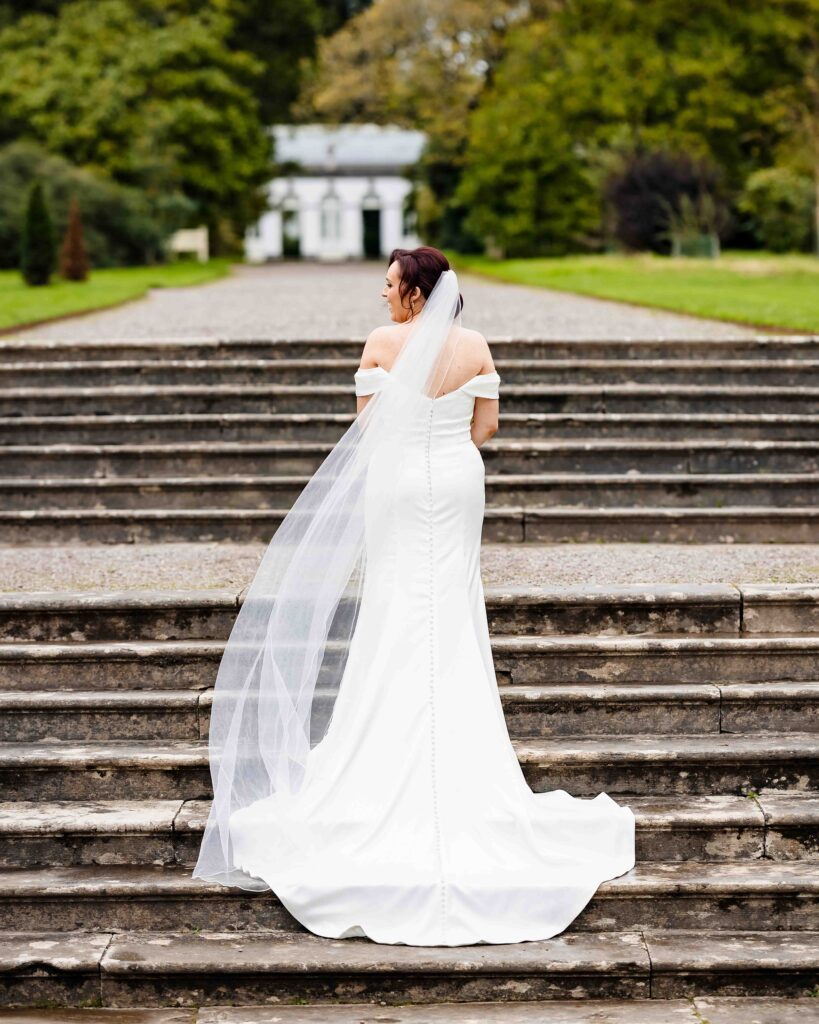Bride seen from behind with her veil trailing, standing on the stone steps in the gardens at Fota House and Gardens, Cork