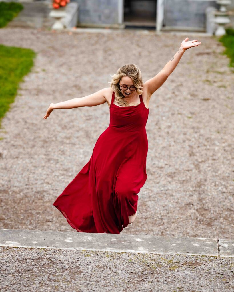 Bridesmaid in a red dress laughing and dancing with her arms out on a gravel path