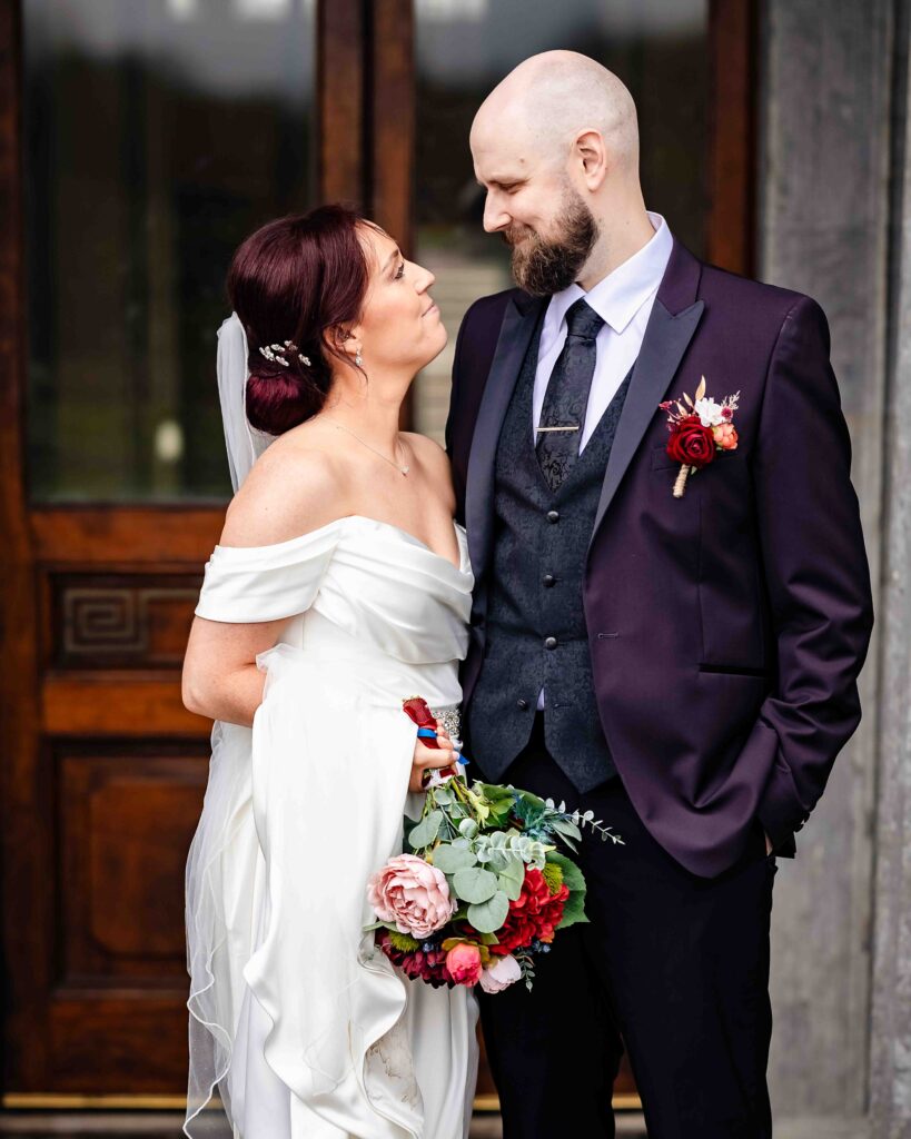 Bride and groom gazing at each other beside a carved wooden door, groom in a purple suit and bride holding a pink and red bouquet