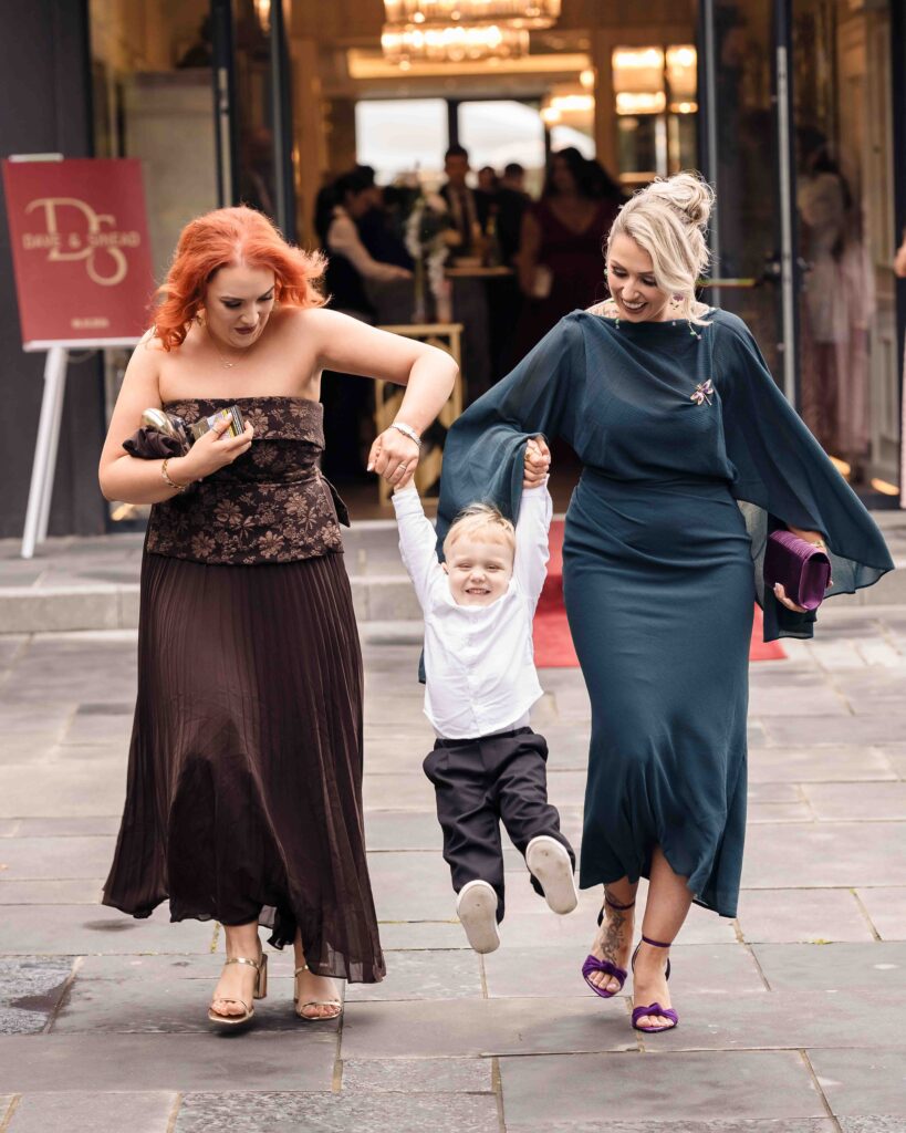 Two women swinging a giggling toddler between them as wedding guests arrive at the reception entrance