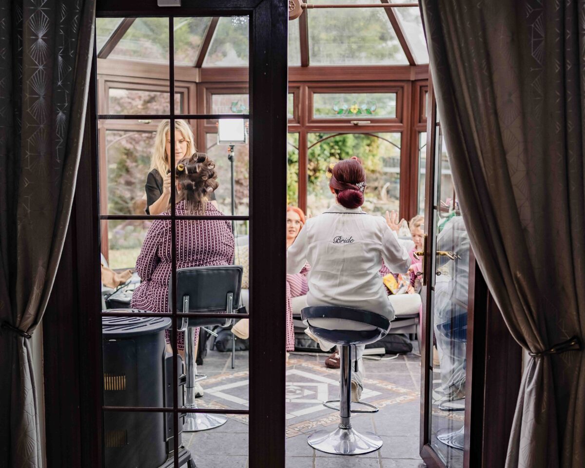 Bridal party having hair and makeup done in a conservatory, seen through glass doors on the wedding morning