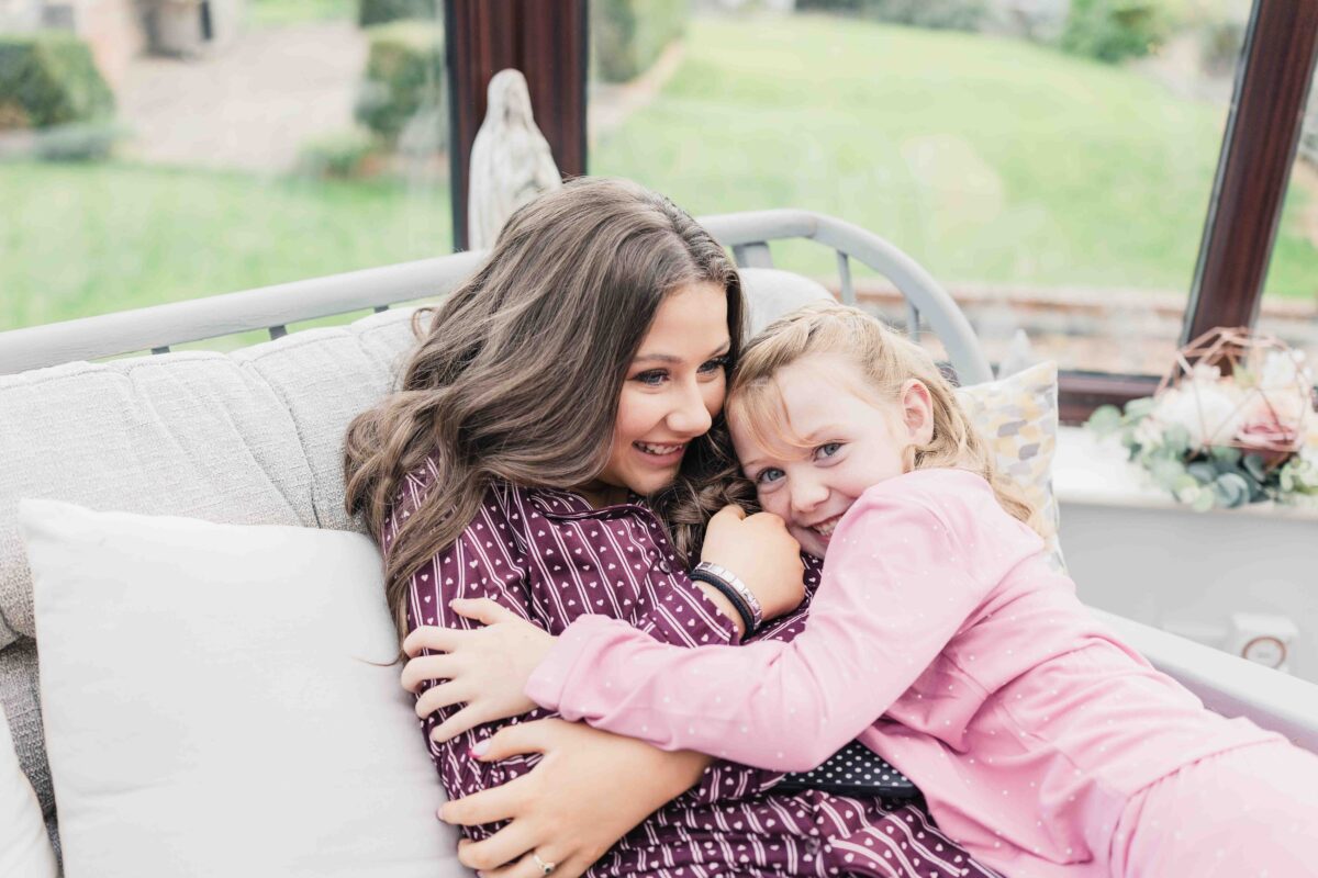 Bridesmaid cuddling a little girl in pink pyjamas on a sofa on the wedding morning