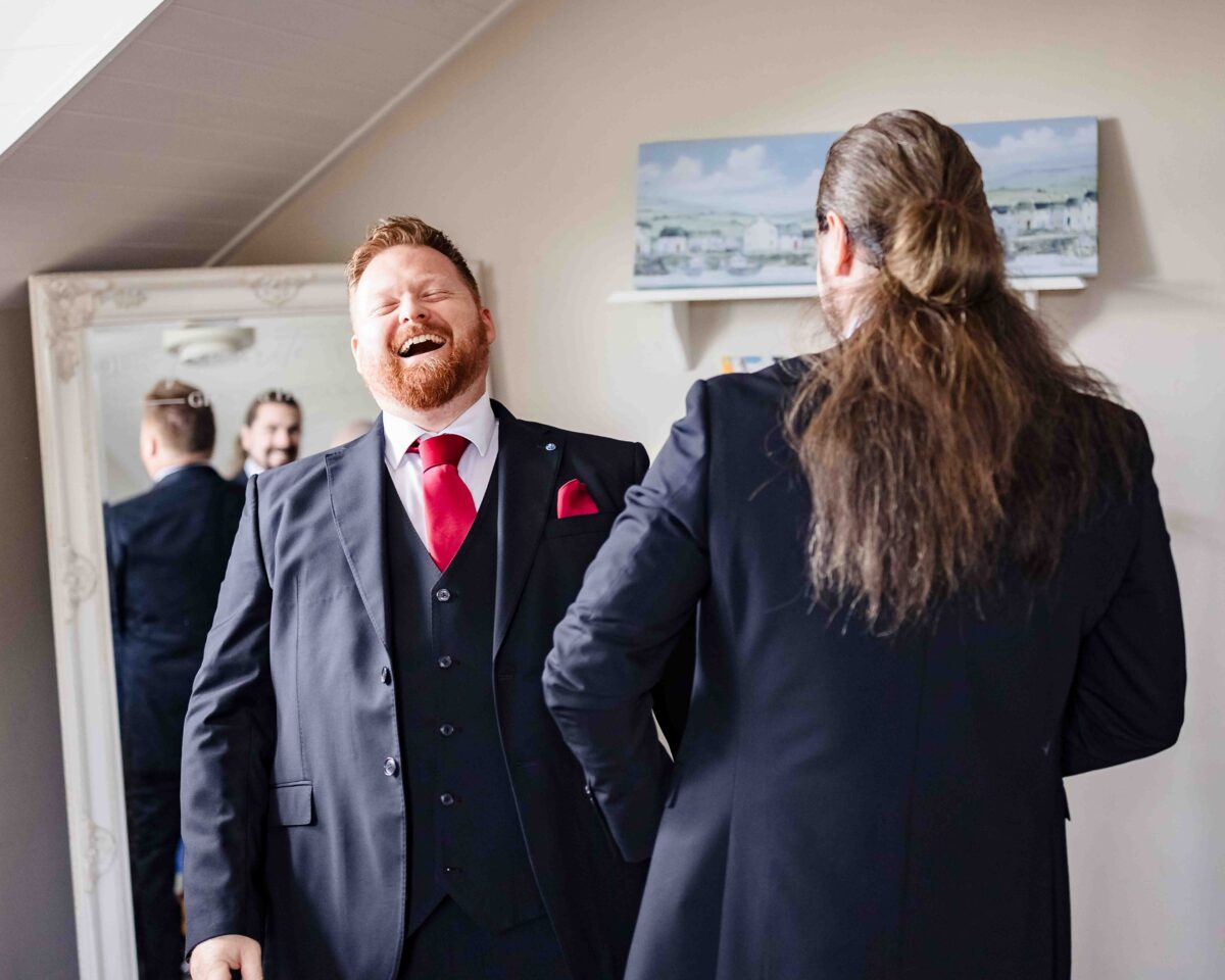 Groomsman laughing in a navy suit and red tie while getting ready beside a large mirror