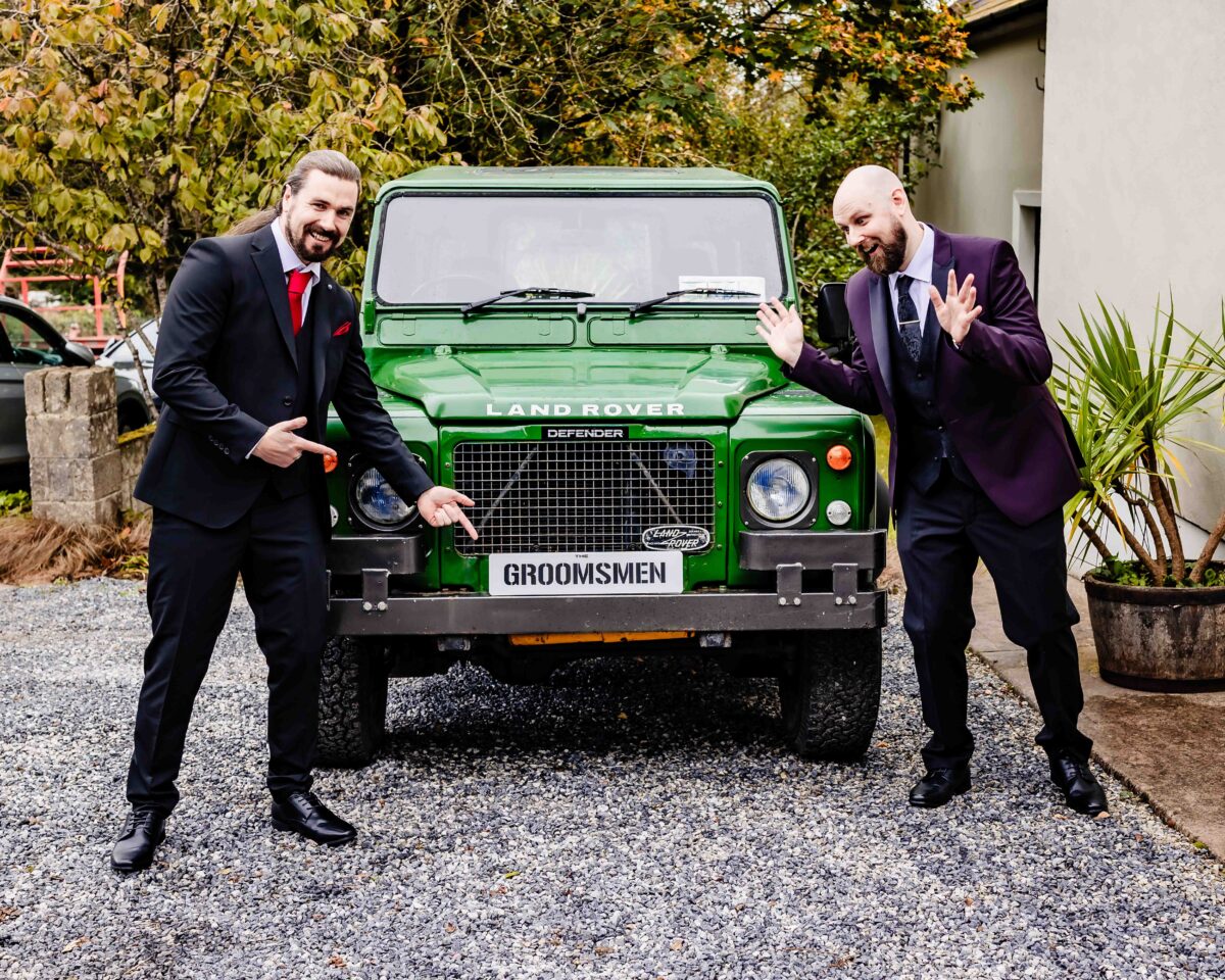 Groom and a groomsman playfully posing beside the green Land Rover Defender with its "Groomsmen" plate
