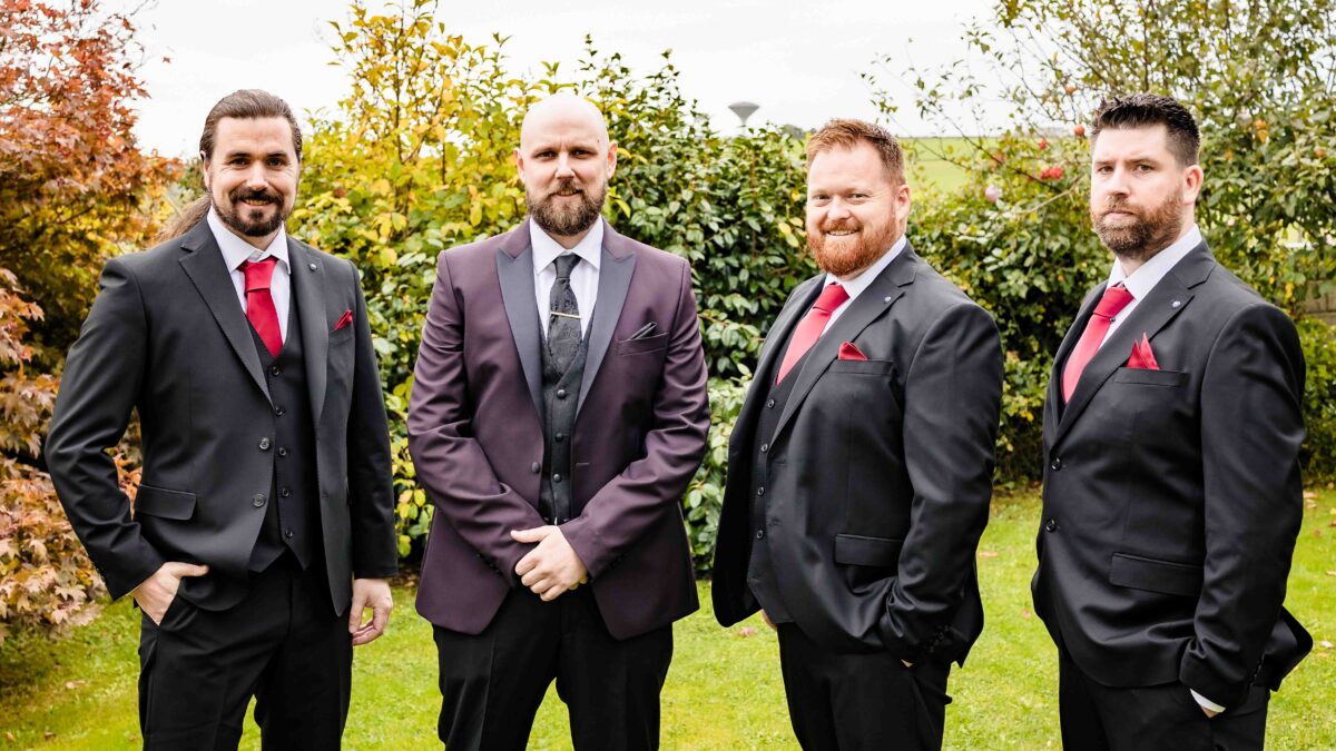 Groom with his three groomsmen in dark suits and red ties standing together in a garden