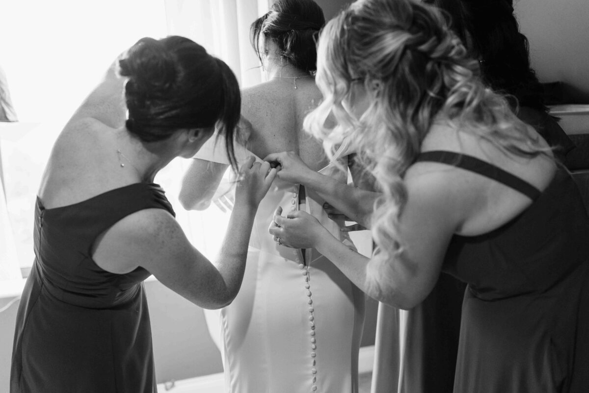 Black and white photo of bridesmaids fastening the buttons on the back of the wedding dress