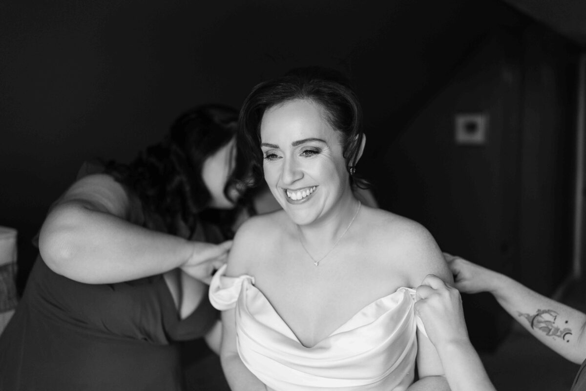 Black and white photo of the smiling bride having her dress adjusted by her bridesmaid