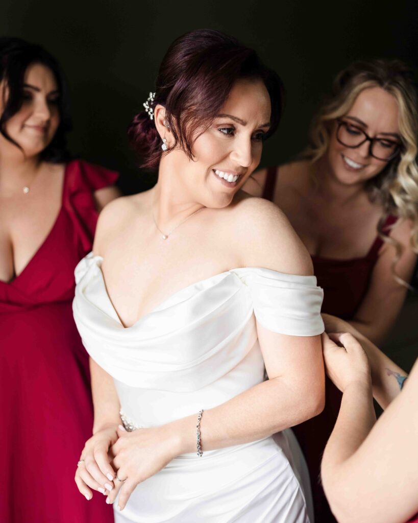 Bride smiling as bridesmaids in red dresses help her into her off-the-shoulder wedding dress