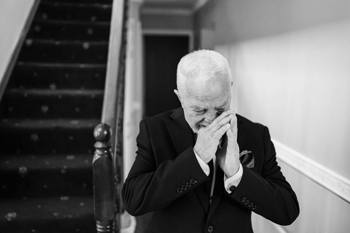 Black and white photo of the bride's father Terry covering his face with his hands at the bottom of the stairs, overcome with emotion