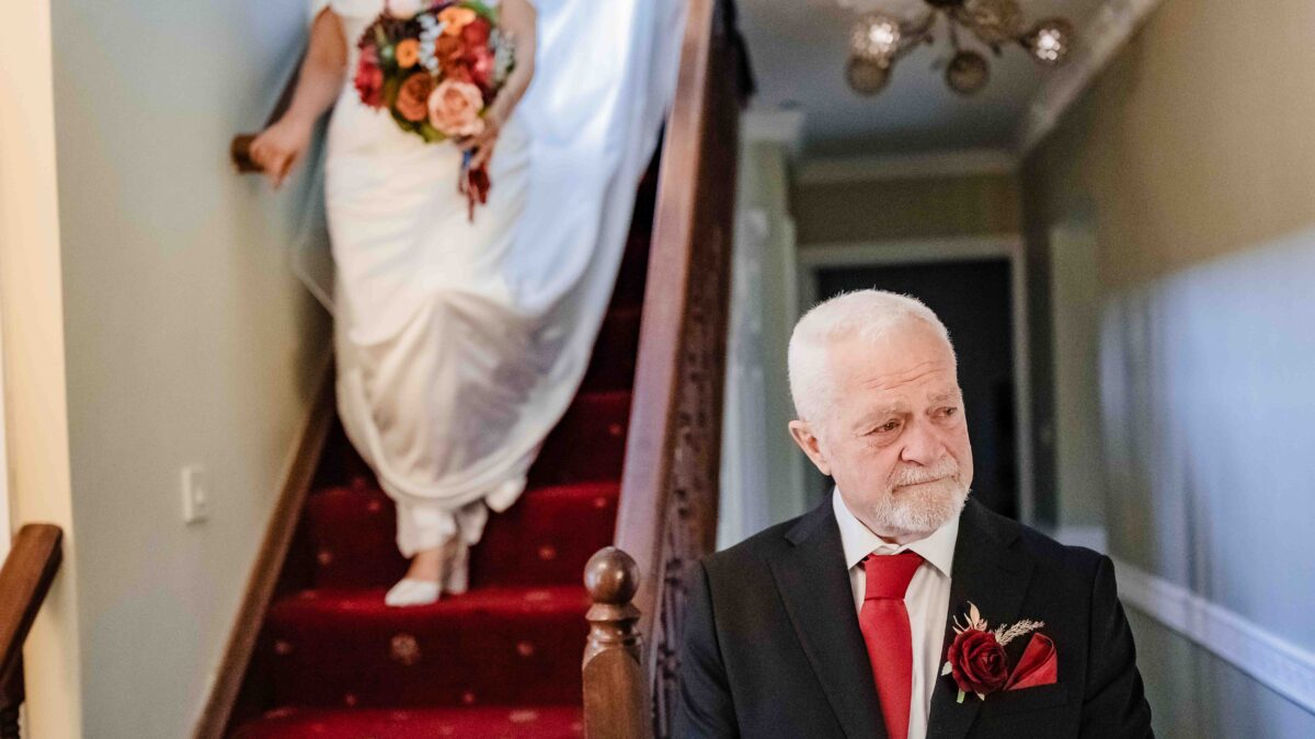 Bride walking down a red-carpeted staircase holding her bouquet as her father Terry looks up at her with emotion