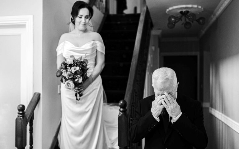 Black and white photo of the bride at the top of the stairs while her father Terry braces himself with his hands over his face