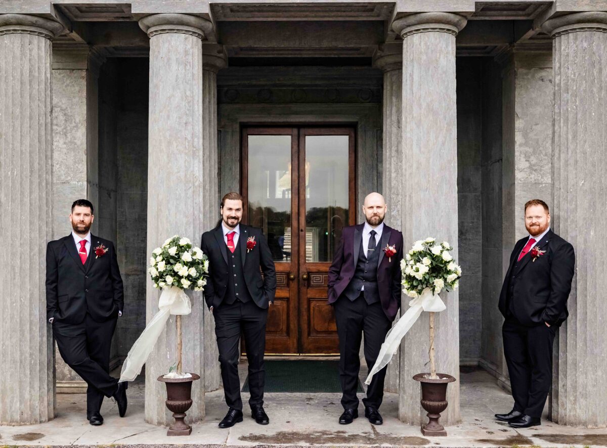 Groom and three groomsmen in dark suits standing between stone columns beside the large wooden front doors of Fota House, Cork
