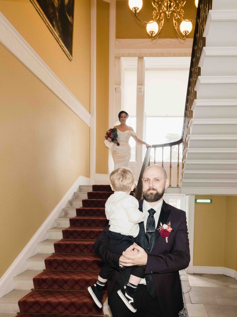 Dave carrying a toddler on the red-carpeted staircase inside Fota House as Sinéad waits at the top, Fota House wedding first look