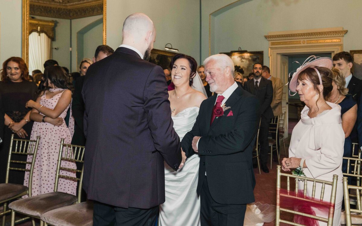Groom greeting the bride's father at the top of the aisle as guests look on in the ceremony room at Fota House, Cork