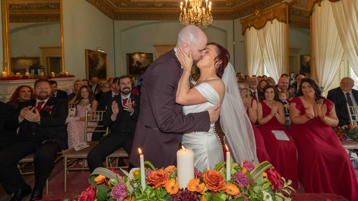 Bride and groom kissing during the ceremony at Fota House and Gardens as guests in red dresses applaud behind an autumn flower arrangement and candles