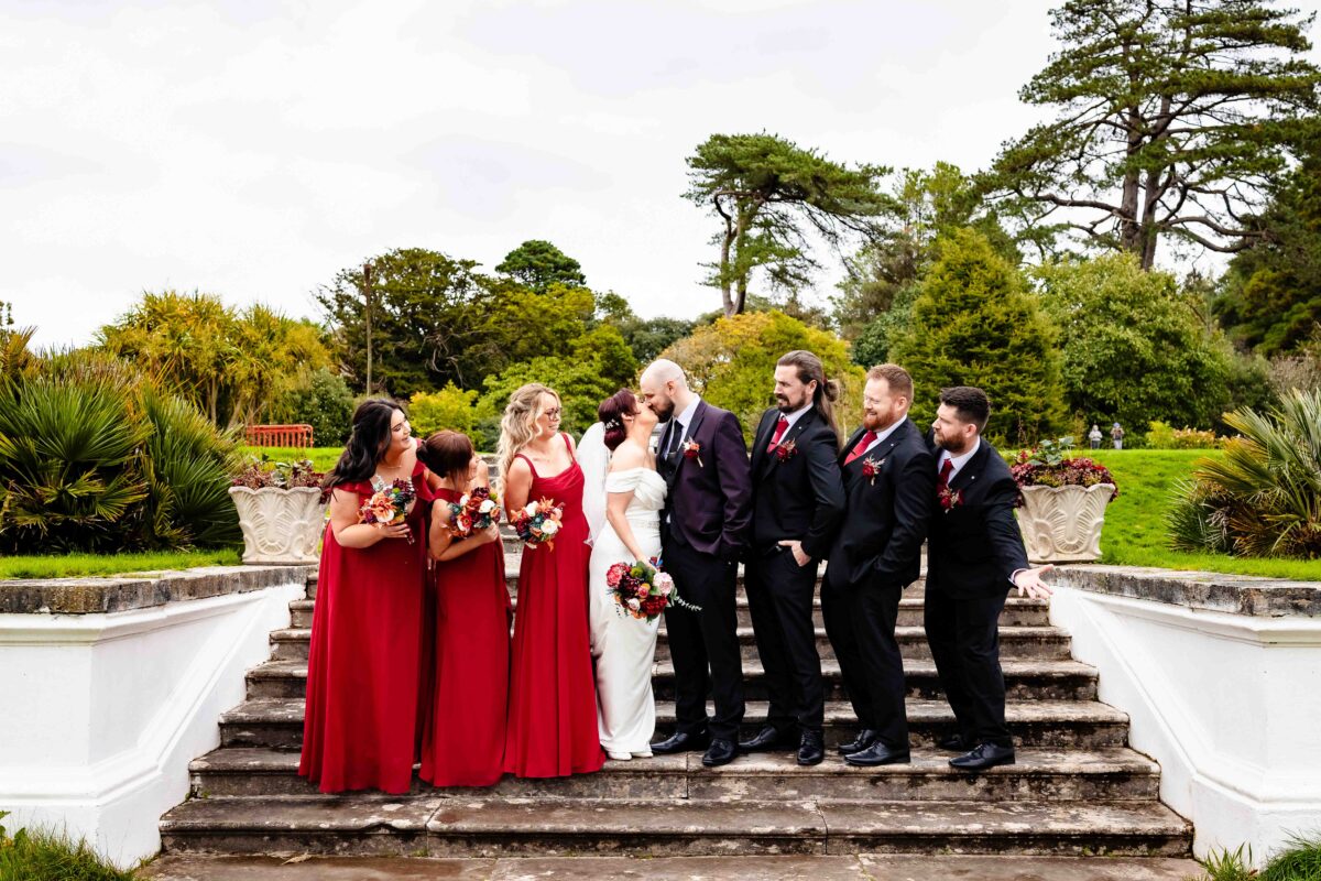 Bridesmaids in red dresses and groomsmen lined up on the stone garden steps at Fota House Gardens as the bride and groom kiss