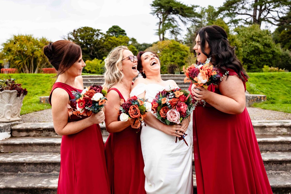 Bride laughing with two bridesmaids in red dresses holding autumn bouquets on stone steps in the gardens at Fota House, Cork