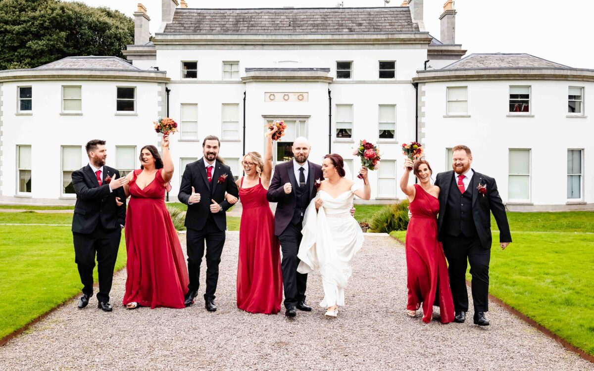 Bridal party walking and cheering toward the camera along the gravel drive in front of Fota House and Gardens, Cork