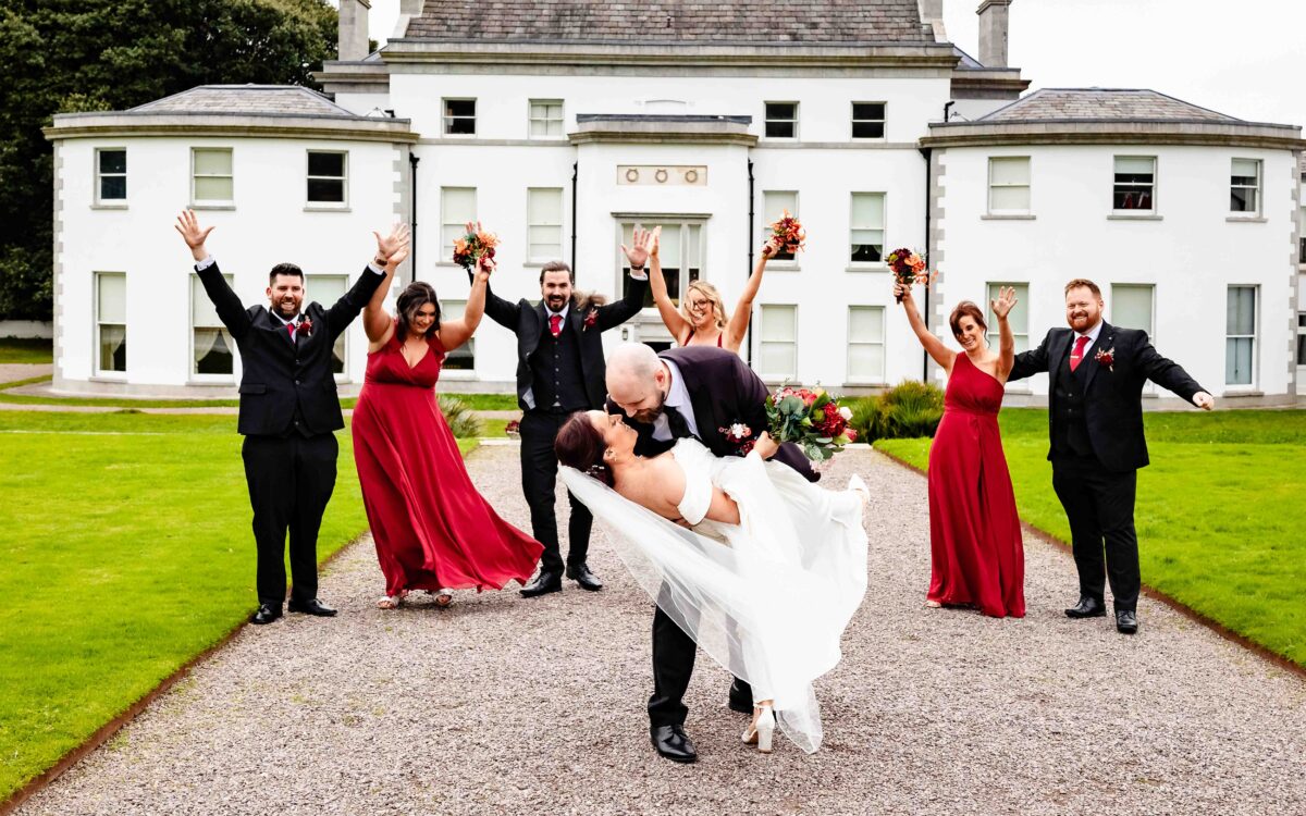 Fota House and Gardens wedding: groom dipping the bride for a kiss as the bridal party cheers on the lawn in front of Fota House, Cork